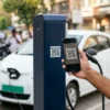 Driver scanning a UBC-enabled EV charging station's QR code with BHIM UPI on a smartphone, with an EV showing a green number plate charging in the background
